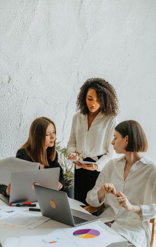 Three women collaborate on a project with charts and laptops in an office.